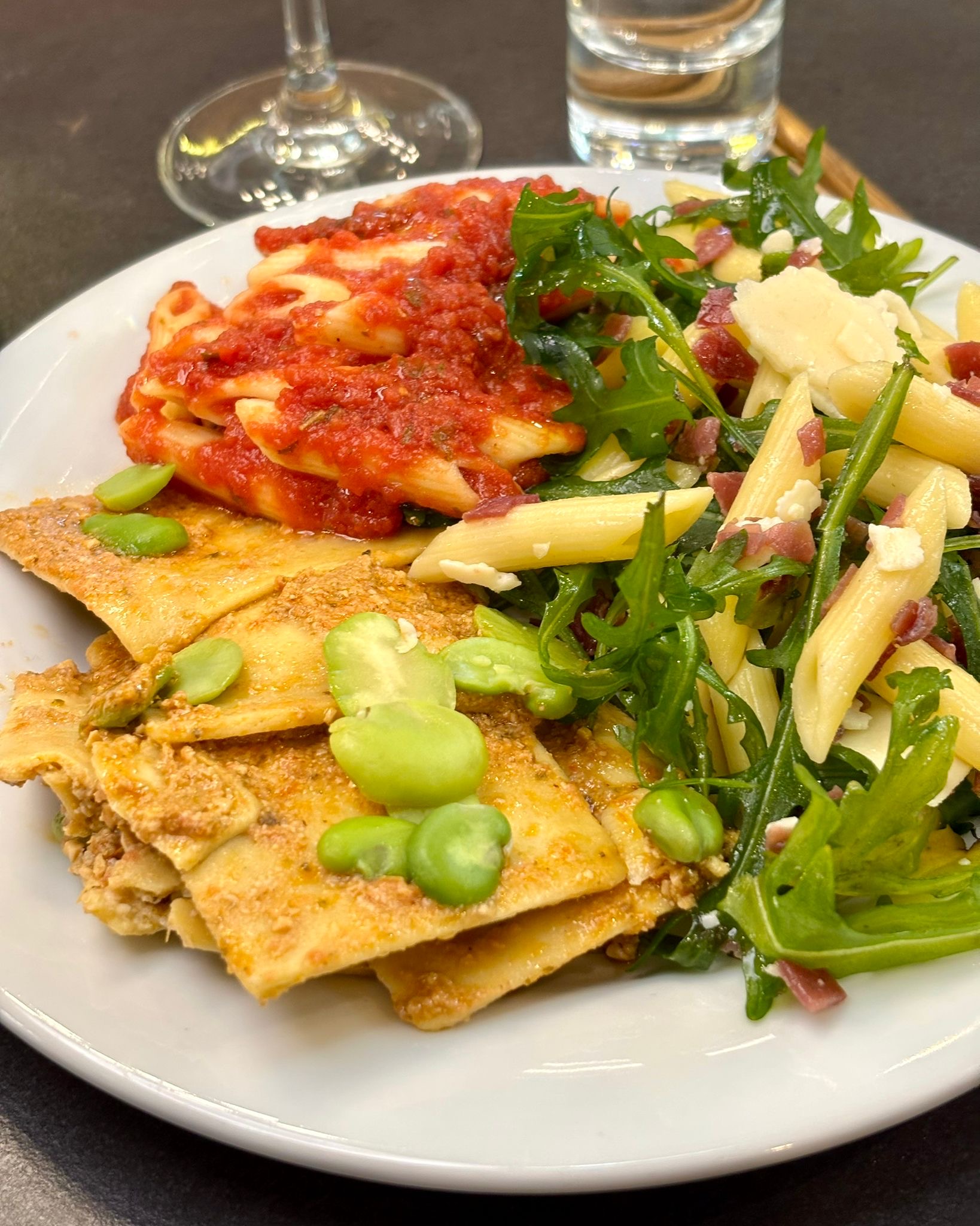Photo montrant un plat typiquement italien - Trio de pâtes composé de raviolis aux artichauts, feves, sauce au pesto rouge - Penne à la bresaola , roquette et parmesan 	- Penne tomates basilique huile d'olive